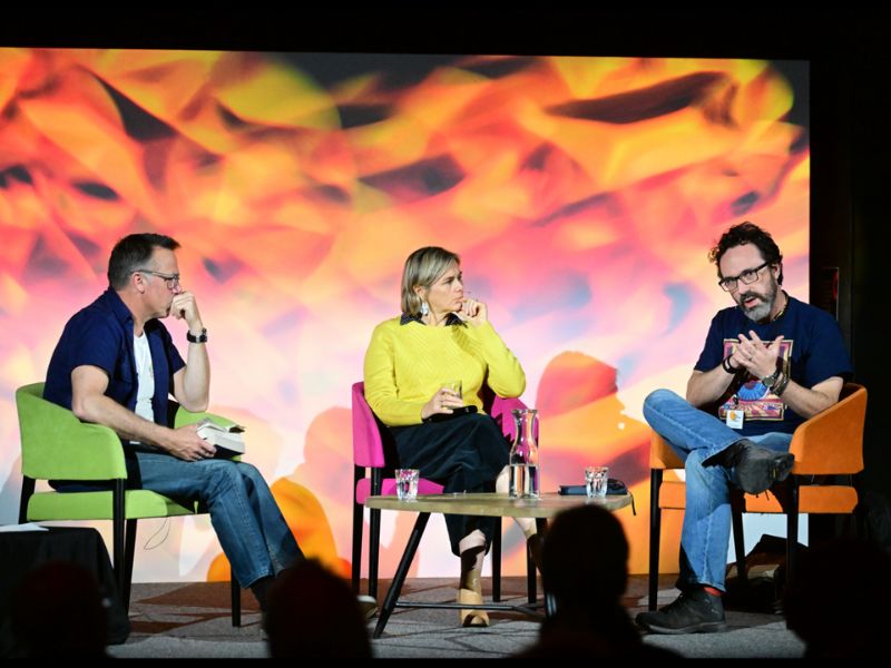 3 people sit on a stage in front of colorful background. 2 are listening intently as one is emphasizing what he is saying with a hand gesture