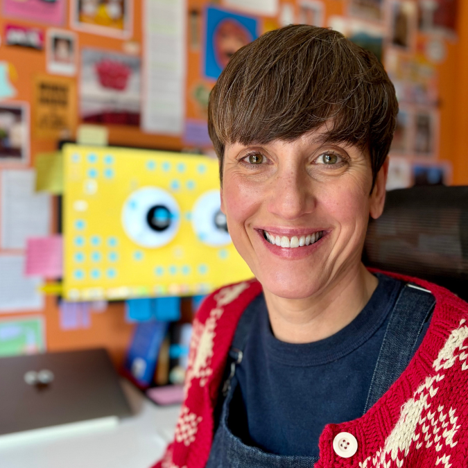 Headshot of author Jo Dabrowski with short brown hair and a fringe, smiling broadly. She is wearing a red and white patterned cardigan over a dark blue top, seated in front of a colourful wall covered in notes and artwork.