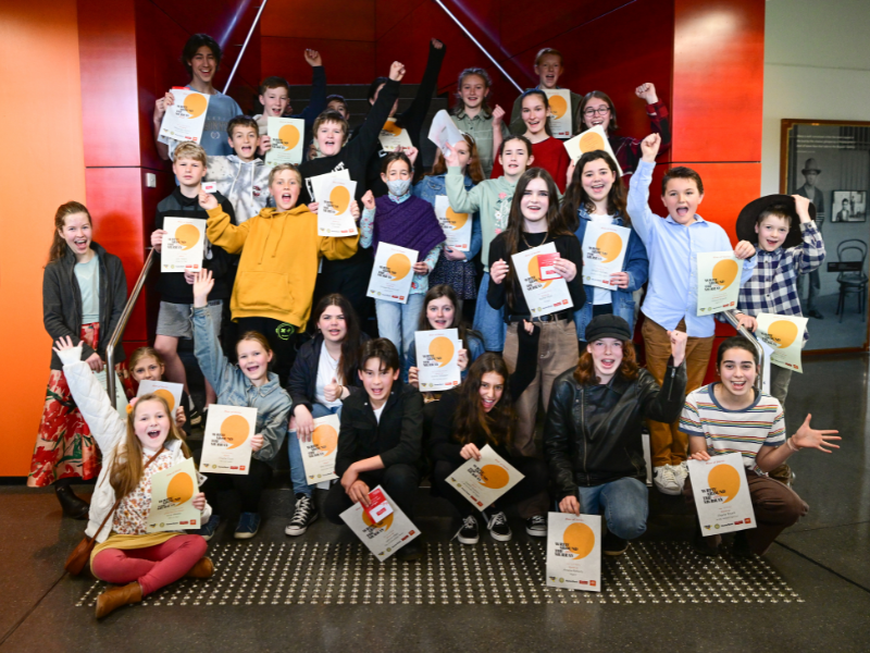 about 30 children grouped together on steps inside the library museum. They are jubilantly cheering and raising their hands in celebration. all are holding River of Stories certificates.