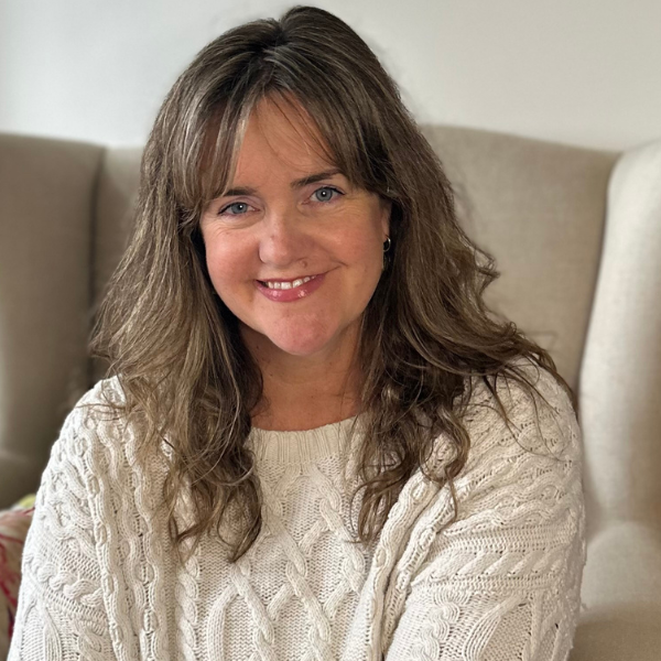 Headshot of author Amy Doak smiling in a cream cable-knit sweater, seated in a beige armchair.