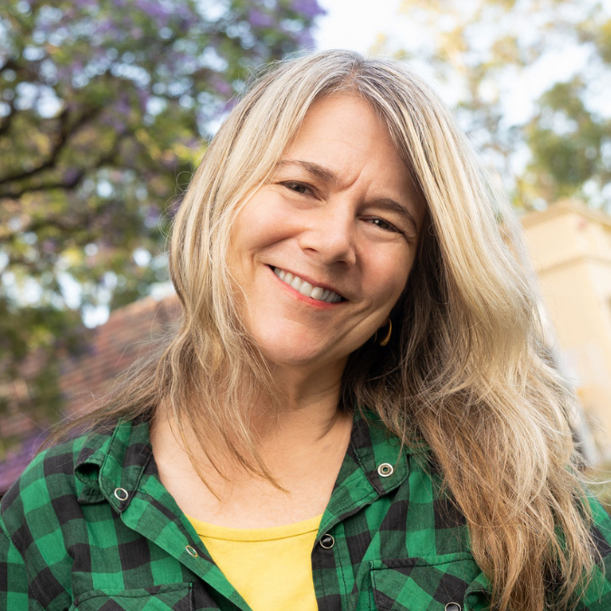 Headshot of author Lucinda Gifford with long blonde hair, smiling outdoors. She is wearing a green and black checkered shirt over a yellow top, with trees in the background.