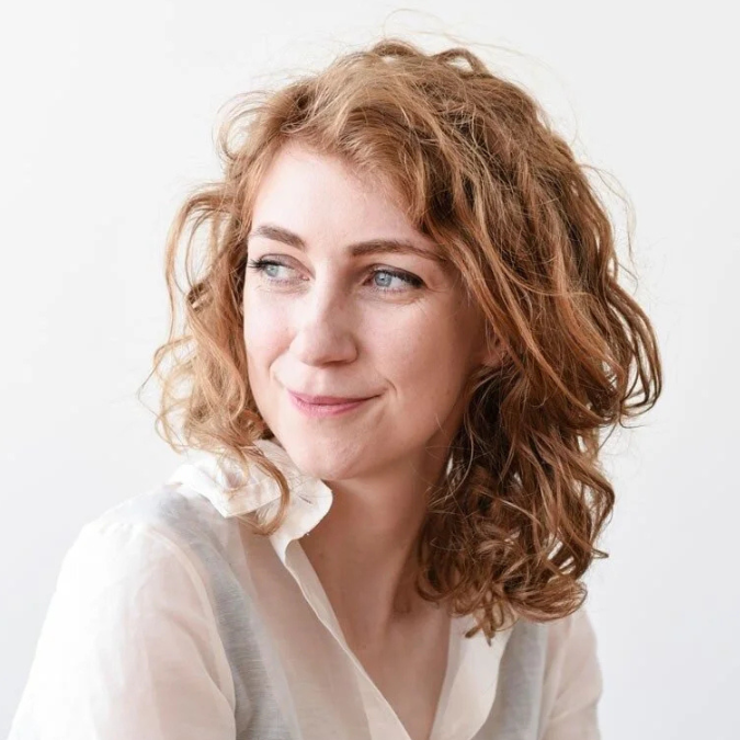 Headshot of author Anna Snoekstra with curly reddish-brown hair, looking to the side with a slight smile against a white background.