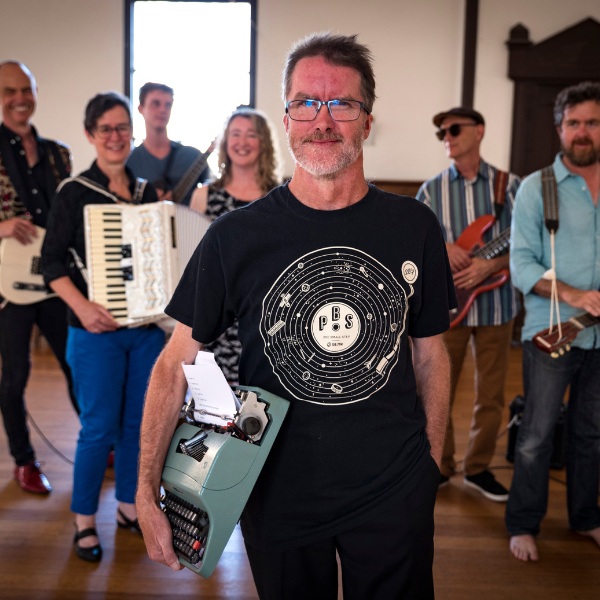 A group of musicians stand smiling each holding a musical instrument whilst the central figure holds a typewriter under his arm